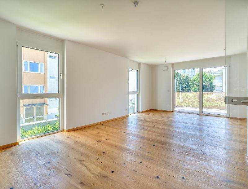 Helles, leeres Wohnzimmer mit Eichenparkettboden und großen Fenstern, die viel Tageslicht hereinlassen.