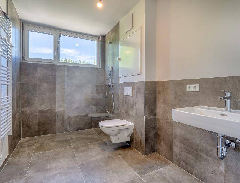 Modernes Badezimmer mit grauen Fliesen, wandhängendem WC und Fenster für Tageslicht.