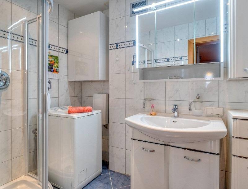Gefliestes Badezimmer mit Duschkabine, Waschbecken, Spiegelschrank und Waschmaschine.