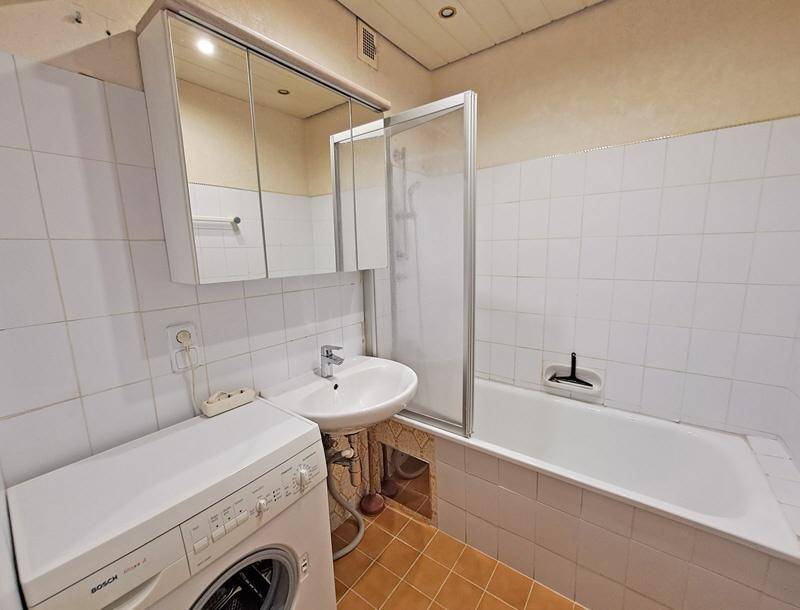 Badezimmer mit weißen Fliesen, Badewanne mit Duschwand, Waschmaschine und Spiegelschrank.