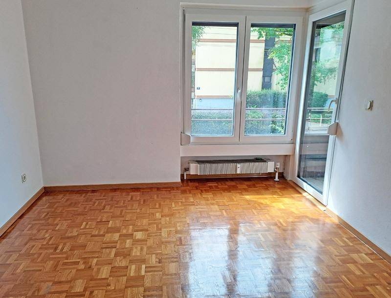 Leeres Zimmer mit glänzendem Parkettboden, großem Fenster und Zugang zu einem Außenbereich.