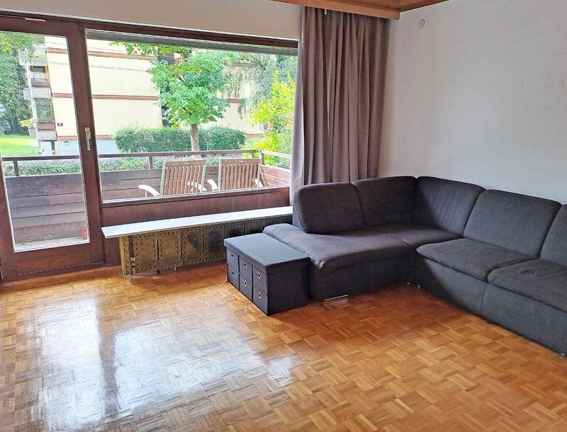 Helles Wohnzimmer mit dunklem Ecksofa, Parkettboden und großem Fenster mit Zugang zum Balkon.