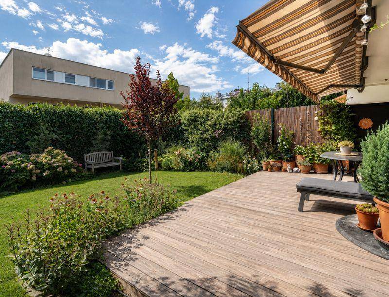 Gepflegter Garten mit Holzterrasse, Rasenfläche, Hecke und ausziehbarer Markise.
