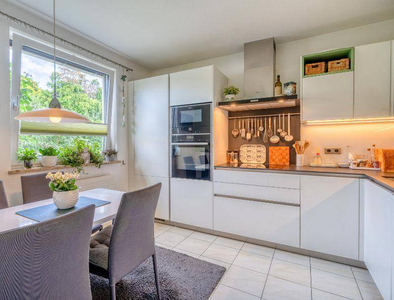 Moderne Einbauküche in Weiß mit integrierten Geräten, Essbereich und großem Fenster.