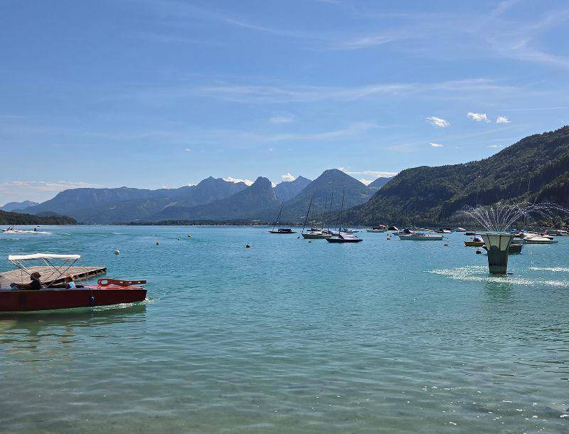 Idyllischer See mit Segelbooten und majestätischen Bergen im Hintergrund unter strahlend blauem Himmel.