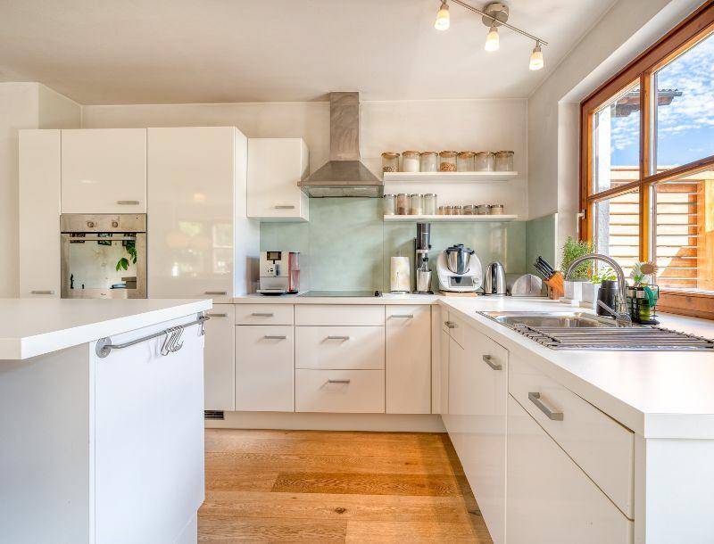 Moderne weiße Einbauküche mit Holzboden, integrierten Geräten und großem Fenster für viel Tageslicht.