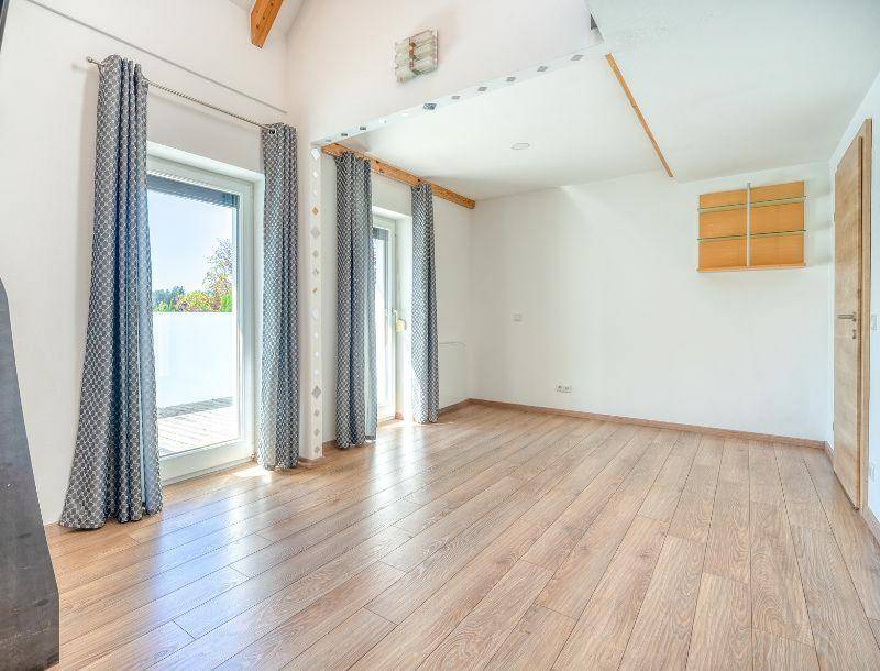 Leerer Raum mit Holzboden, hohen Decken, sichtbaren Holzbalken und Zugang zu einem Balkon.