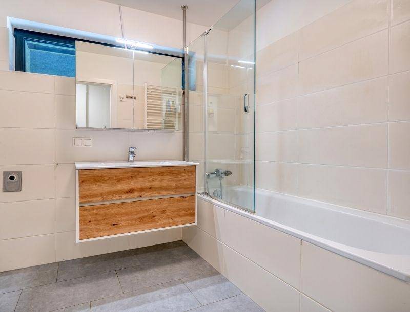 Modernes Badezimmer mit Badewanne, Duschwand, Holzwaschtisch und hellen Fliesen.