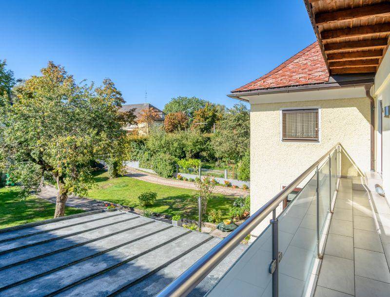 Langer Balkon mit Glasgeländer und Fliesenboden, bietet einen schönen Blick auf den grünen Garten.