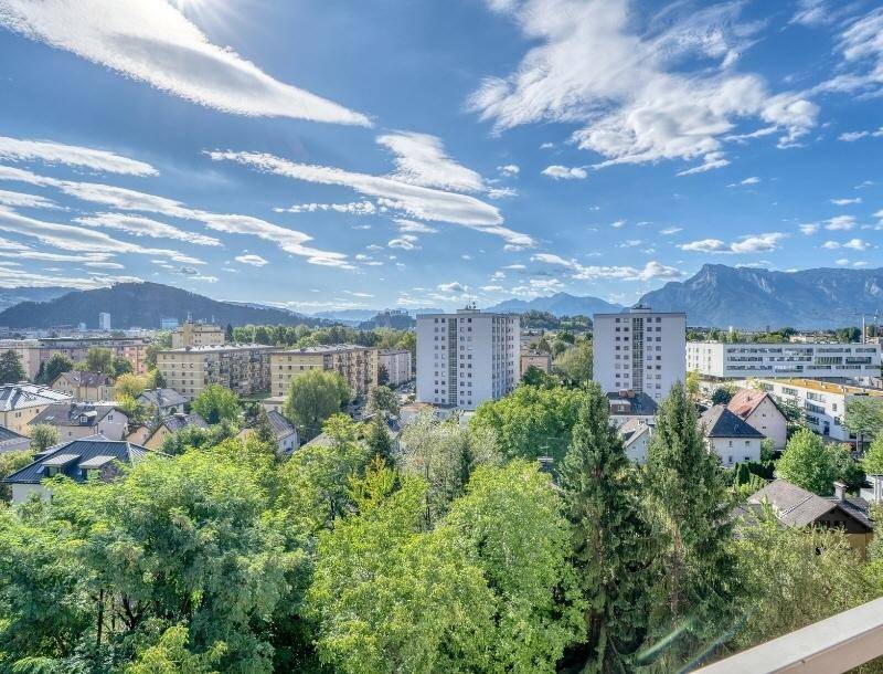 Weitblick über die Stadtlandschaft mit grünen Bäumen und Bergen im Hintergrund unter blauem Himmel.