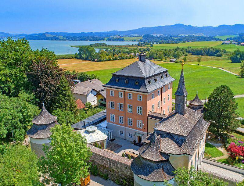 Historisches Schlossgebäude mit Türmen und einem See im Hintergrund, eingebettet in eine malerische Landschaft.