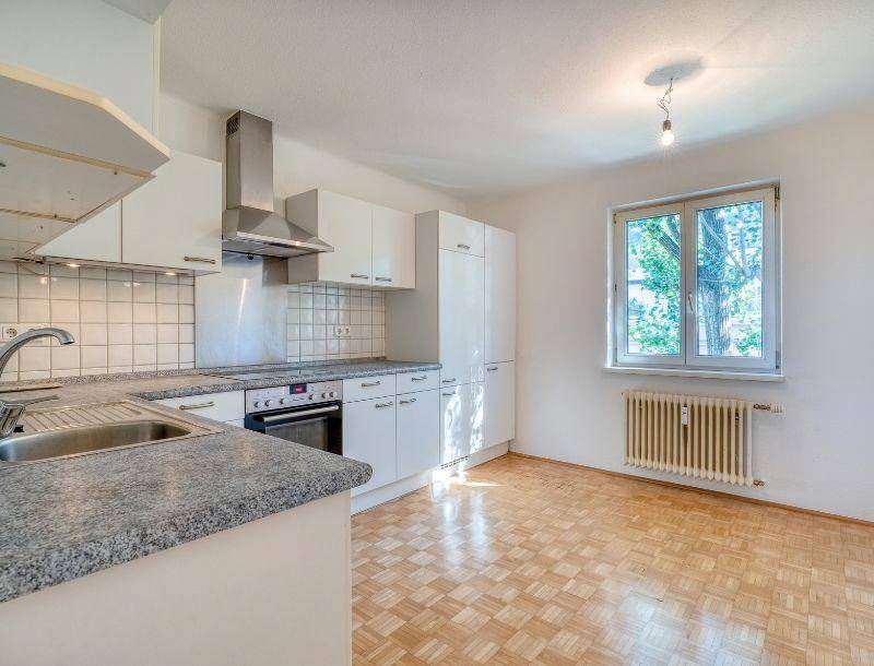 Funktionale Küche mit weißen Schränken, grauer Arbeitsplatte und Parkettboden, gut beleuchtet durch ein Fenster.