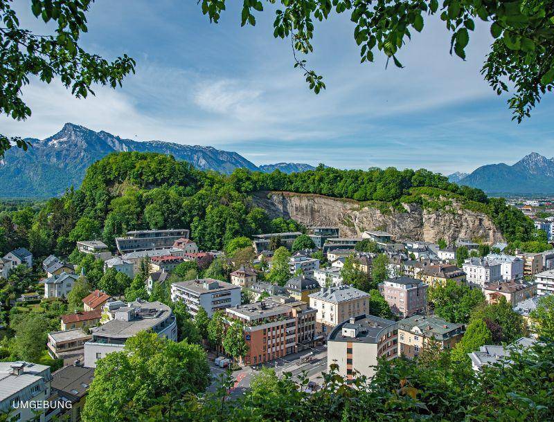 Panoramablick auf eine Stadtlandschaft umgeben von grünen Hügeln und majestätischen Bergen unter blauem Himmel.