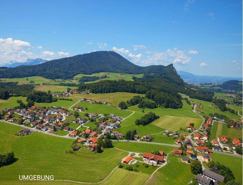 Weitläufige Landschaftsaufnahme mit ländlicher Siedlung, grünen Feldern und Bergen im Hintergrund.