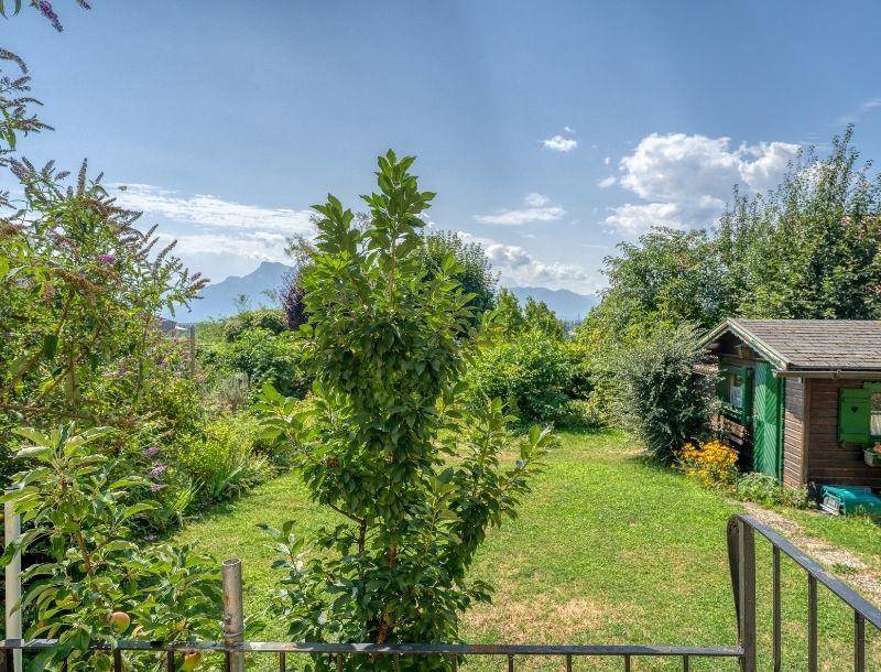 Grüner Gartenbereich mit Bäumen, Sträuchern und einem kleinen Holzschuppen mit Bergblick.