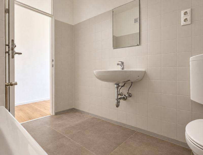 Badezimmer mit hellen Fliesen, Waschbecken, Toilette und Zugang zu einem weiteren Raum.