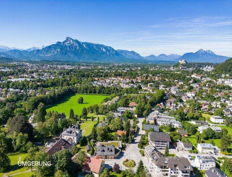 Panoramablick aus der Vogelperspektive auf eine Stadtlandschaft mit viel Grün und majestätischen Bergen im Hintergrund.