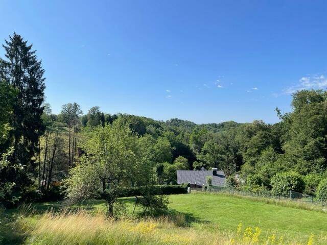Panoramablick über eine weite grüne Landschaft mit bewaldeten Hügeln und einem Haus unter blauem Himmel.