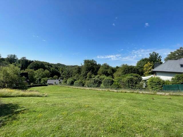 Sonnige Wiesenfläche mit Blick auf bewaldete Hügel und ein Haus am rechten Bildrand.