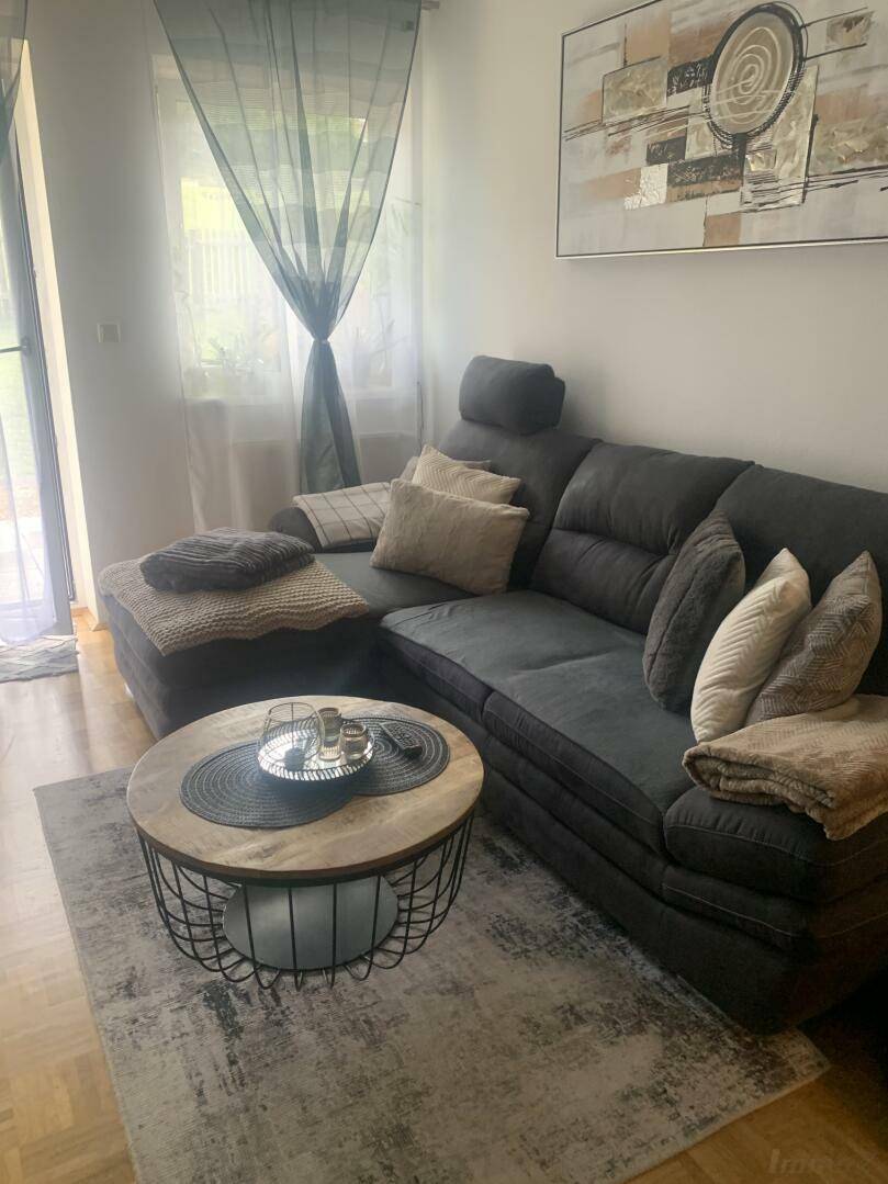 Gemütliches Wohnzimmer mit dunklem Ecksofa, Couchtisch und Teppich, hell durch Fenster.