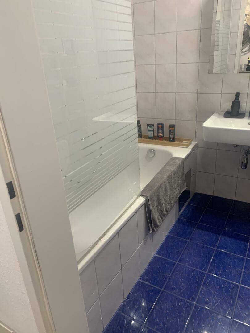 Badezimmer mit blauen Bodenfliesen, weißer Badewanne und Waschbecken, funktional eingerichtet.