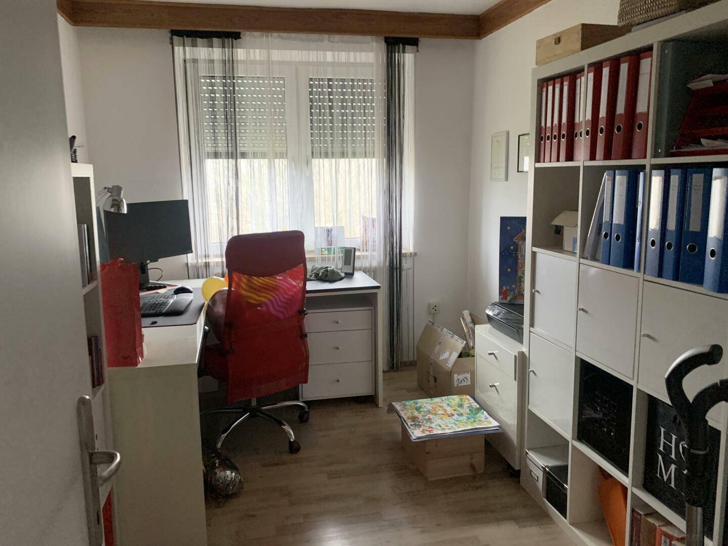 Arbeitszimmer mit Schreibtisch, Bürostuhl und Regalen, gut beleuchtet durch ein Fenster.