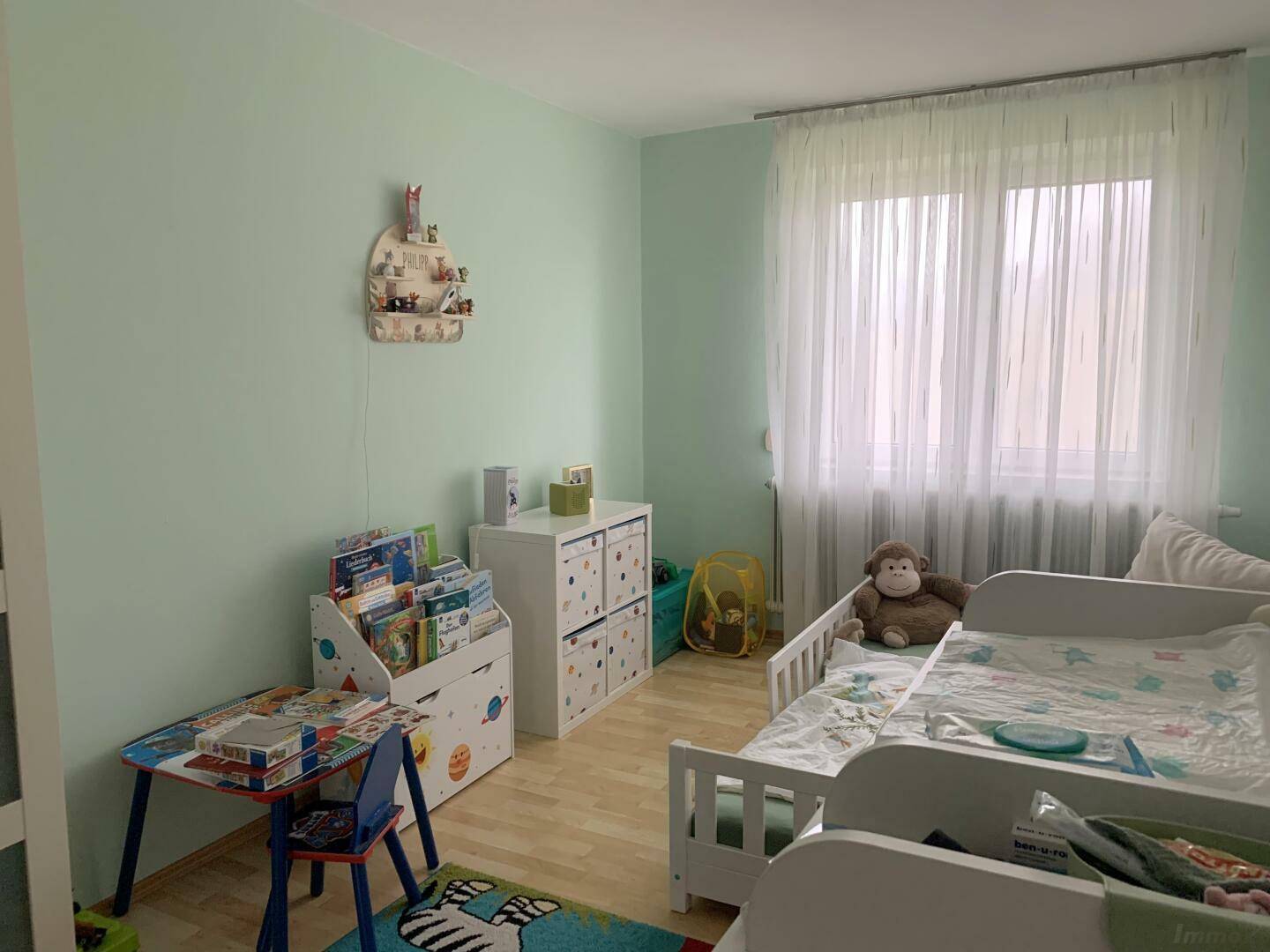 Kinderzimmer mit hellgrünen Wänden, Holzboden und Spielzeug, gut beleuchtet durch ein Fenster.