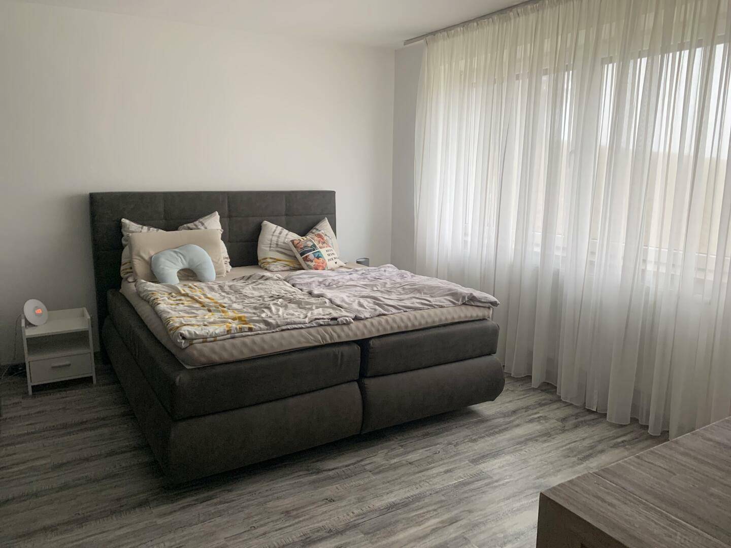 Helles Schlafzimmer mit Doppelbett, Laminatboden und großen Fenstern mit Vorhängen.