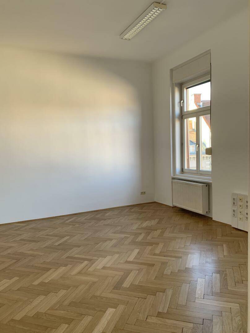 Leeres Zimmer mit Fischgrätparkett, einem hohen Fenster und weiß gestrichenen Wänden.