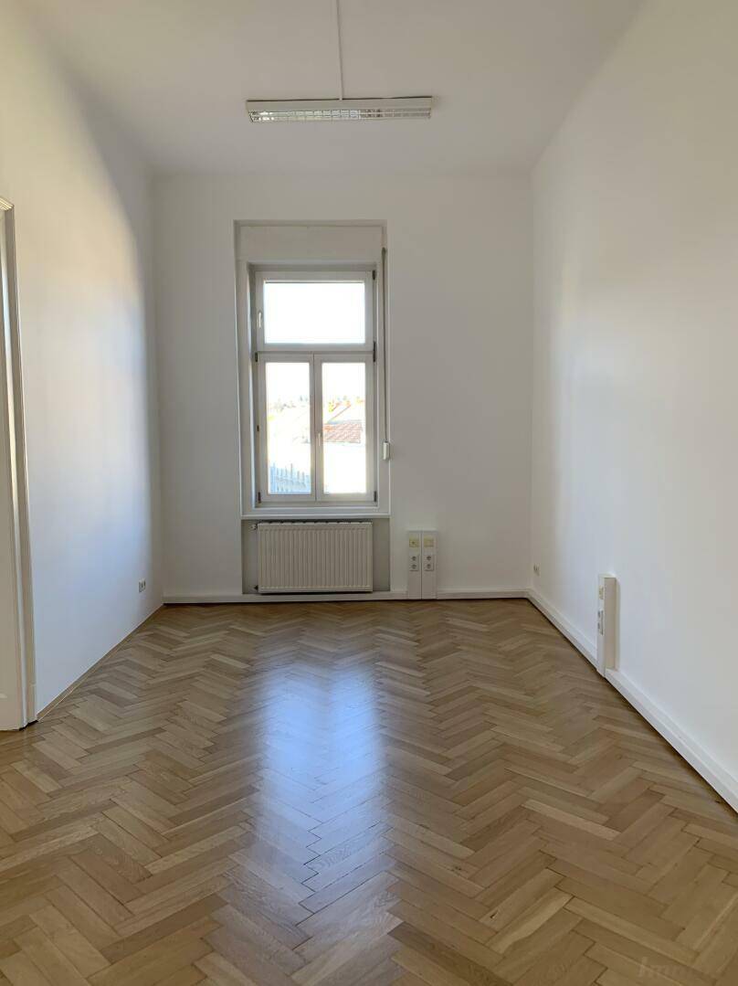 Langes, helles Zimmer mit Fischgrätparkett, einem hohen Fenster und weiß gestrichenen Wänden.