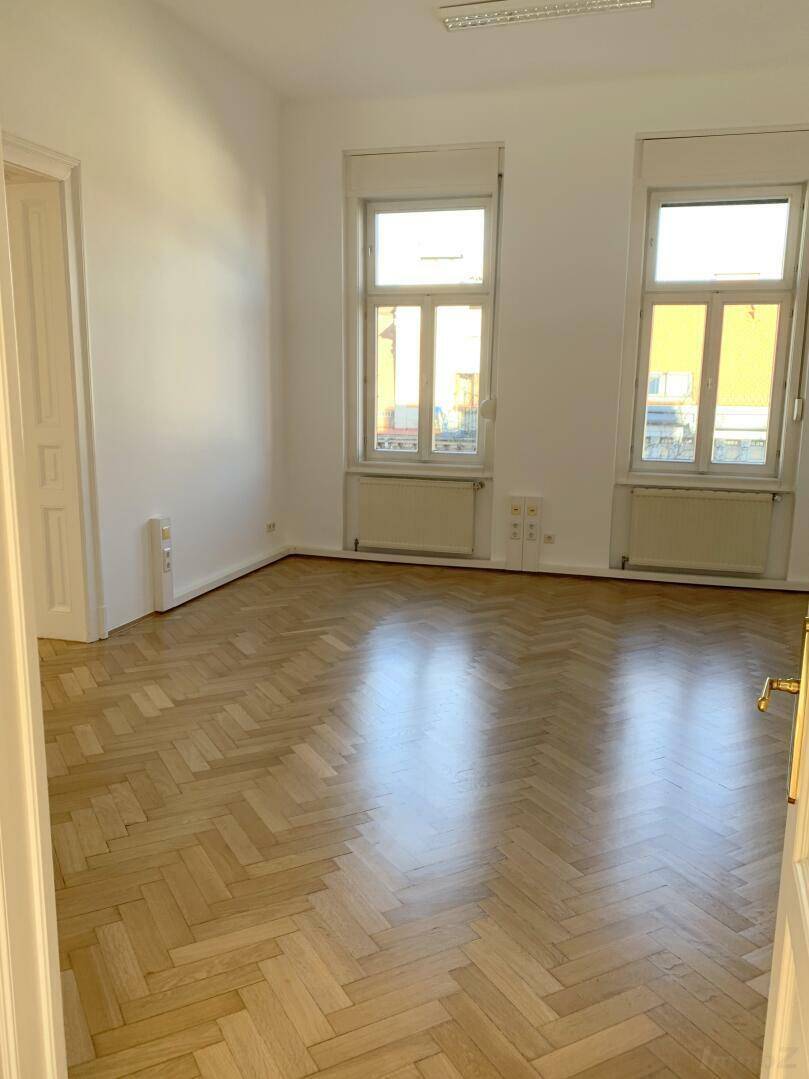 Heller Raum mit glänzendem Fischgrätparkett, hohen Fenstern und weiß gestrichenen Wänden.