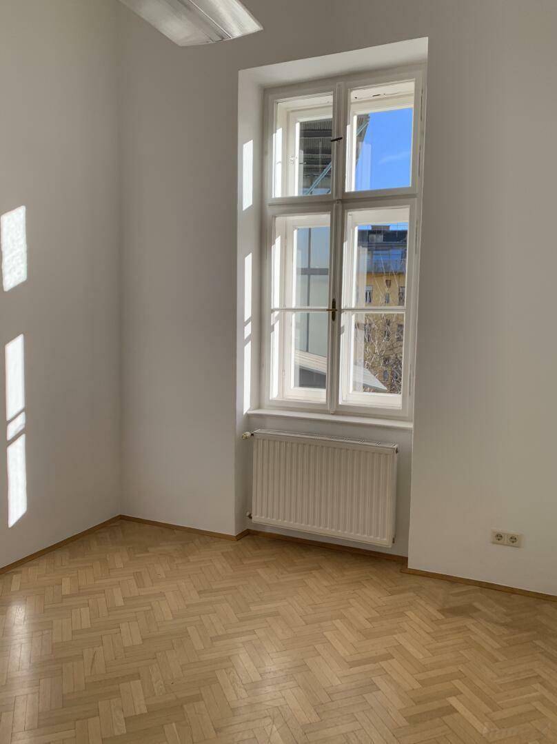 Helles Zimmer mit Fischgrätparkett, großem Fenster und Heizkörper unter dem Fenster.