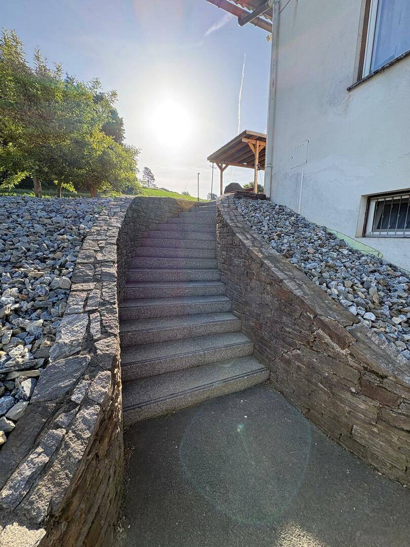 Steintreppe mit Trockenmauern führt zu einem höher gelegenen Bereich mit Pergola im Sonnenlicht.