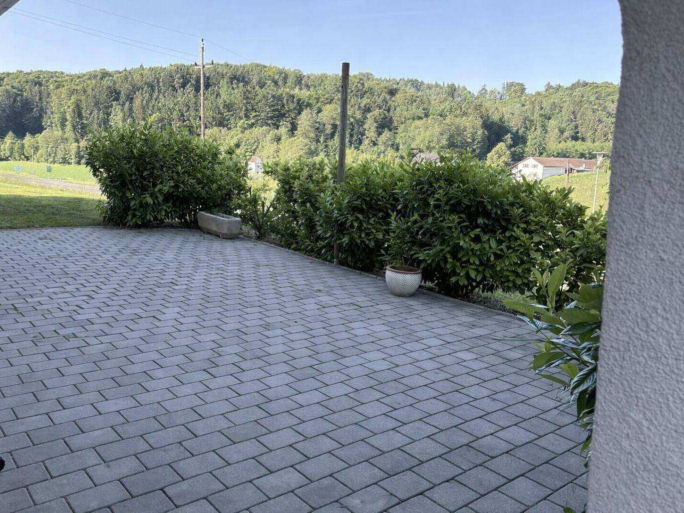 Gepflasterte Terrasse mit Hecken und weitem Blick auf die umliegende grüne Hügellandschaft.
