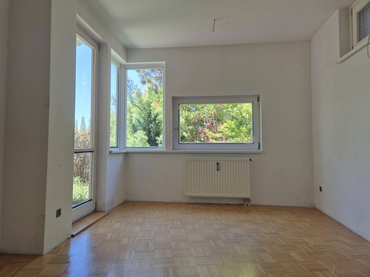 Heller Raum mit Parkettboden und großen Fenstern, die viel Tageslicht und Grünblick bieten.