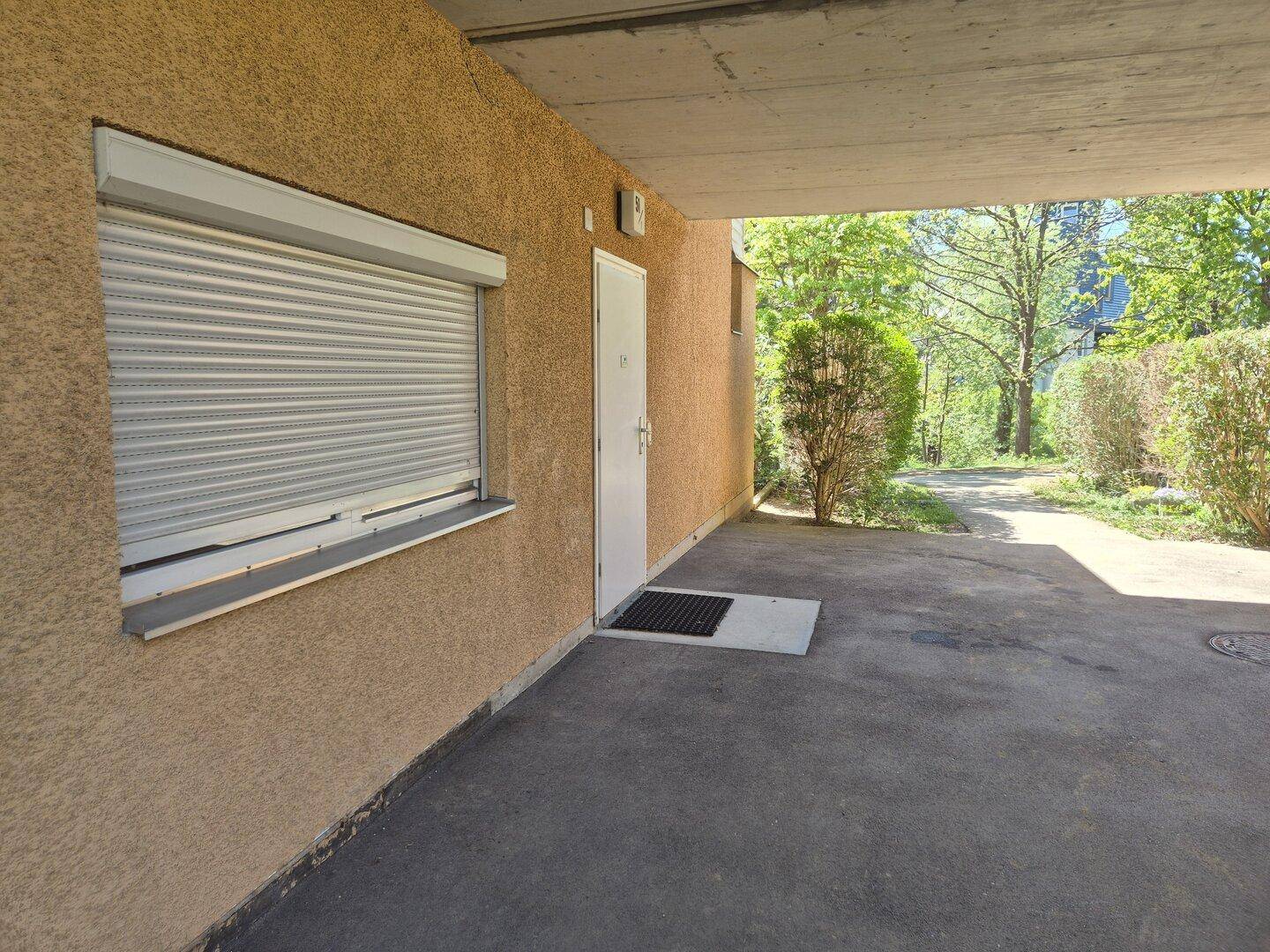 Eingangsbereich eines Gebäudes mit Rollladenfenster und überdachtem Zugangsbereich im Freien.