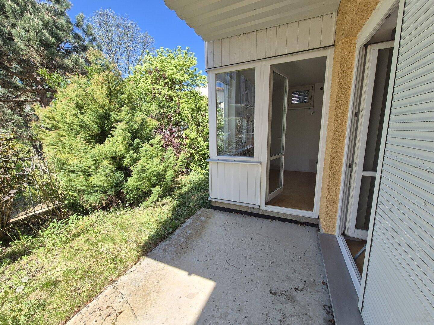 Kleine Terrasse mit Zugang zu einem verglasten Wintergarten, umgeben von üppigem Grün.
