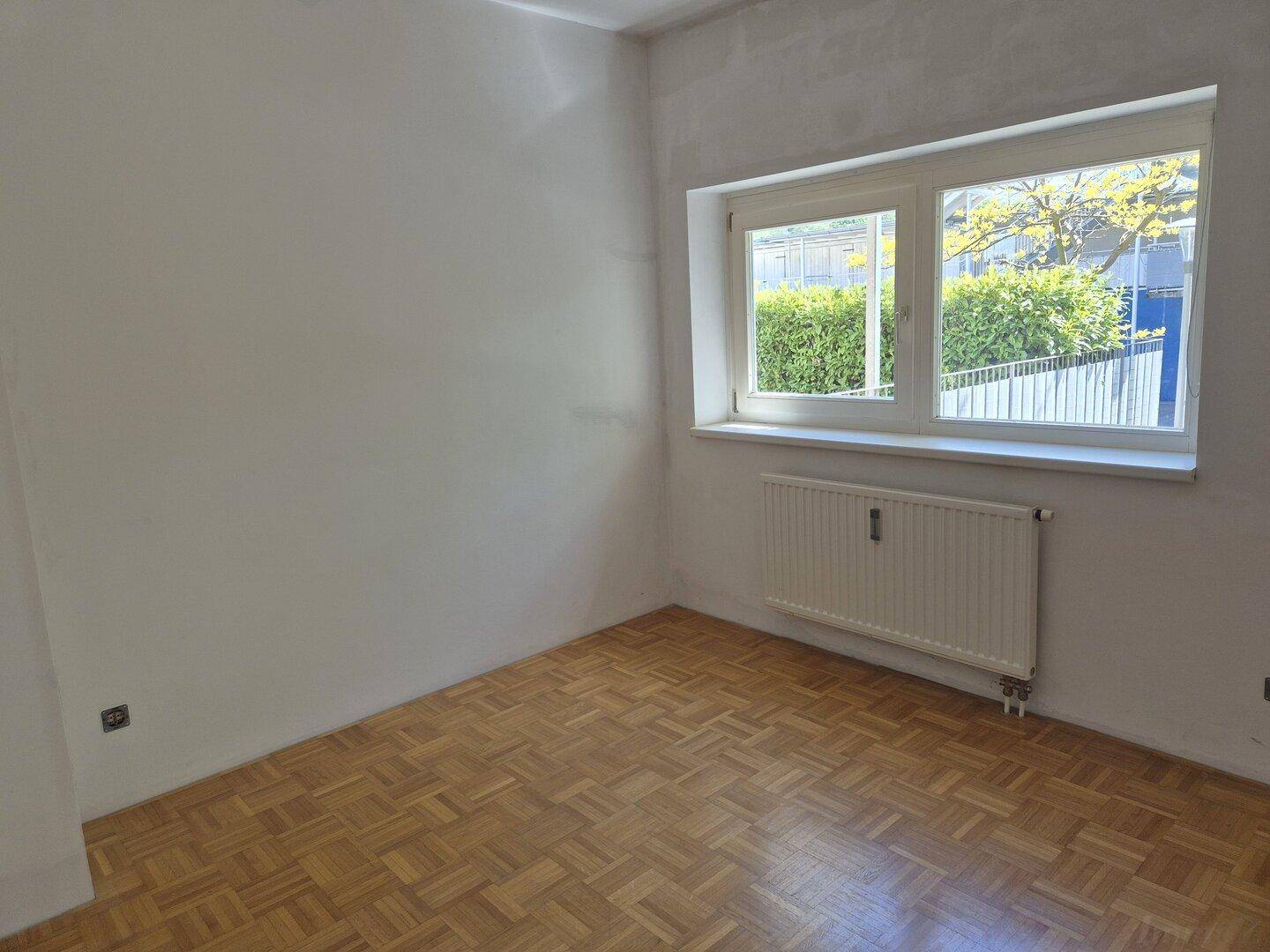 Helles Schlafzimmer mit Parkettboden und Fenster mit Blick ins Grüne.