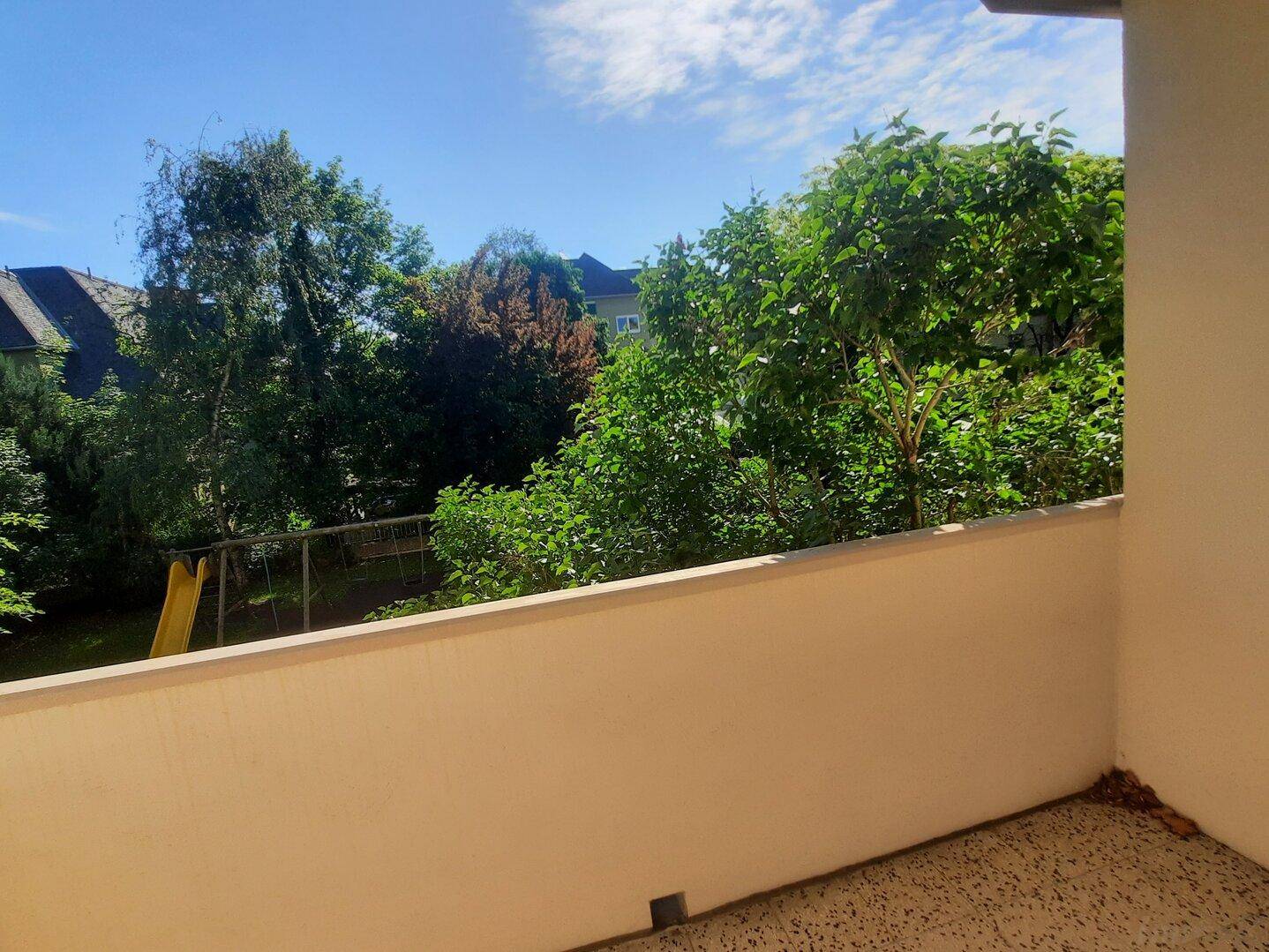 Blick von einem Balkon ins Grüne mit Bäumen und einem Spielplatz im Hintergrund.