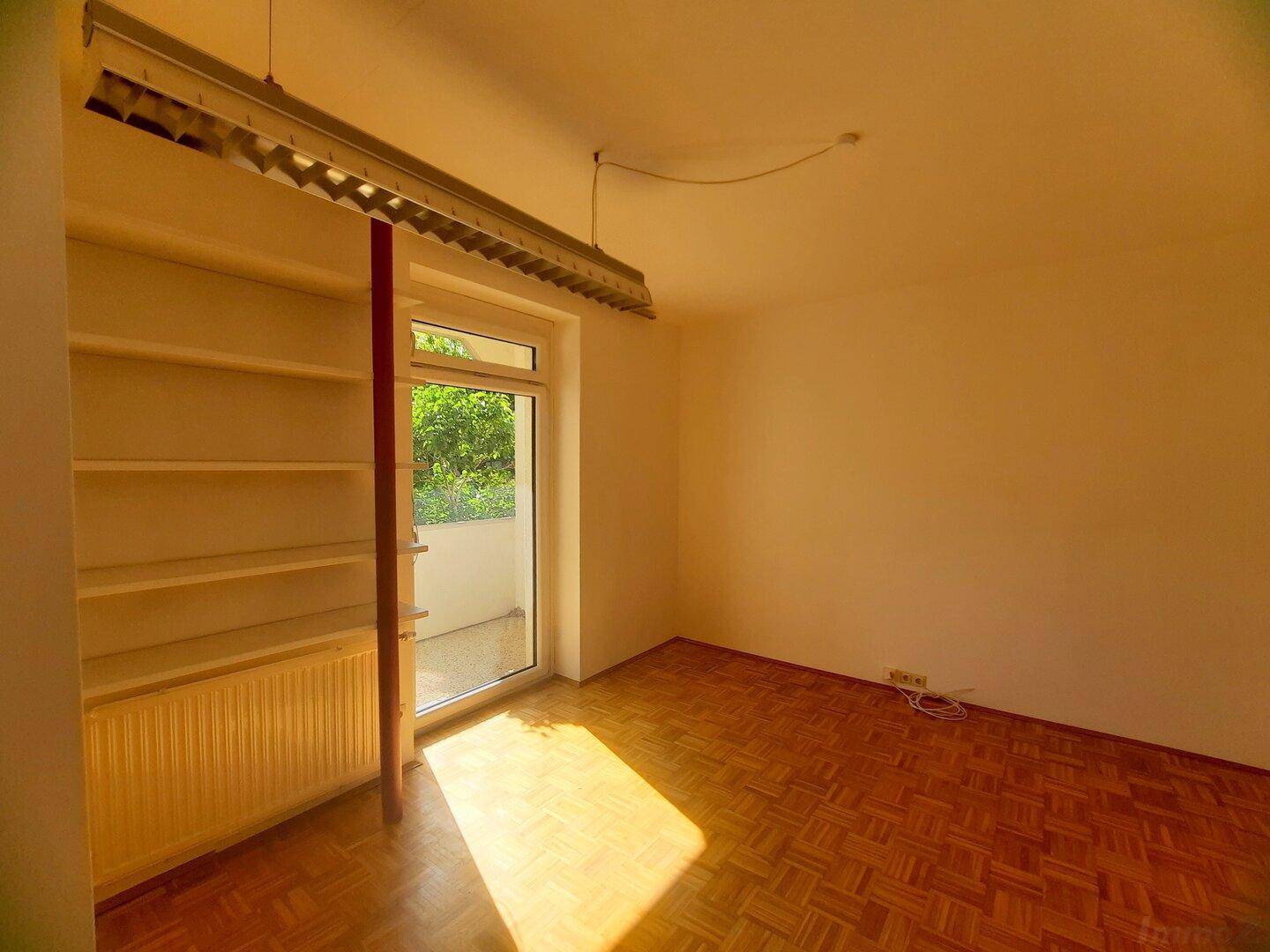 Helles Zimmer mit Parkettboden, Heizkörper und Zugang zu einem kleinen Balkon.