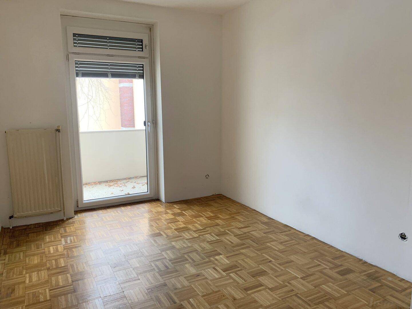 Leeres Zimmer mit Parkettboden, Rollladen und Zugang zu einem Außenbereich.