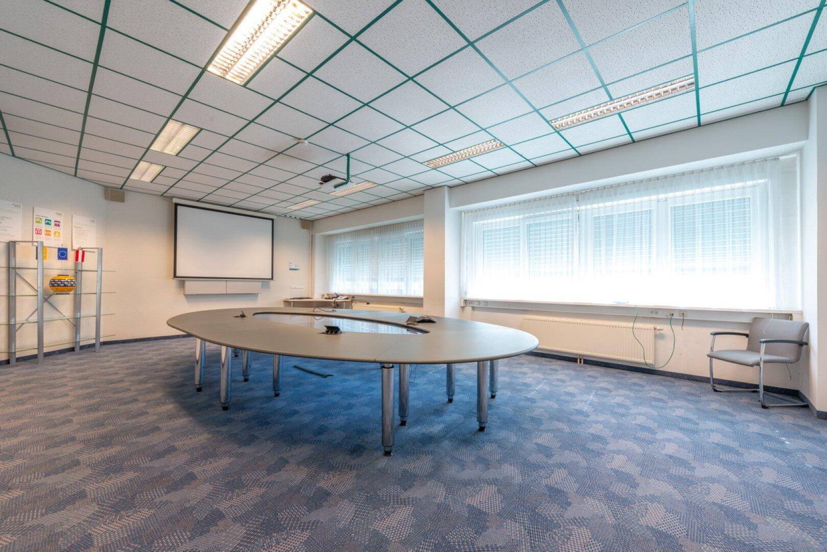 Besprechungsraum mit großem ovalen Konferenztisch, Projektionsfläche und Teppichboden.