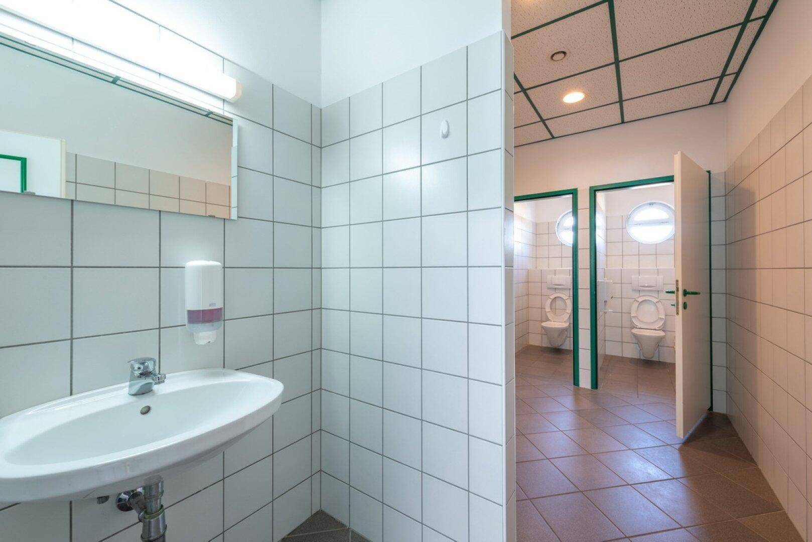 Heller Sanitärbereich mit weißen Fliesen, Waschbecken und zwei Toilettenkabinen.