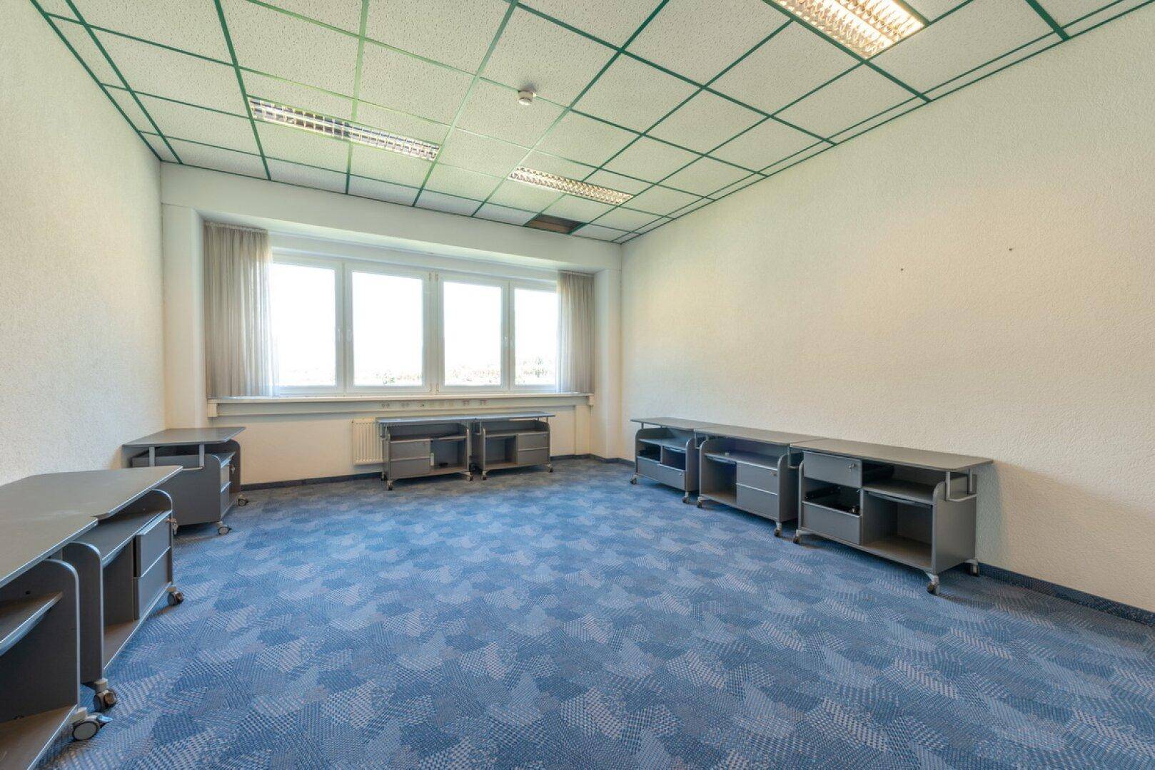 Leerer Büroraum mit blauer Teppichboden und mobilen grauen Schränken unter großen Fenstern.