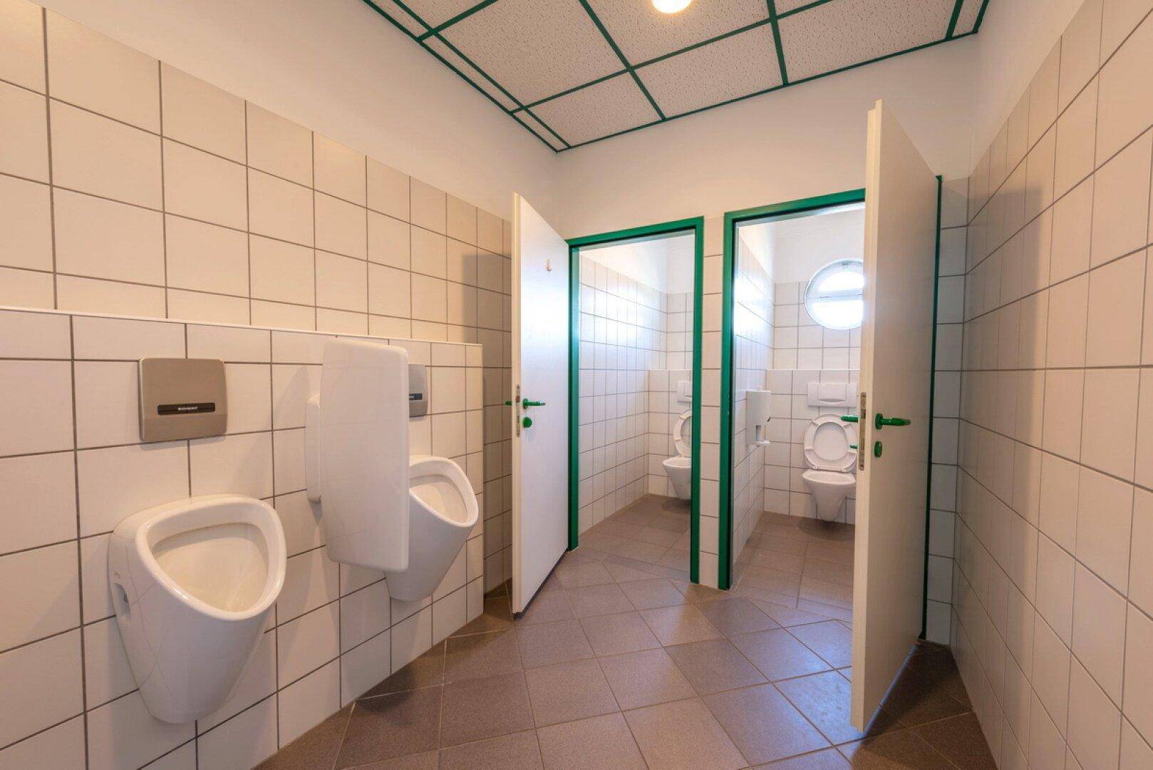 Gepflegter Sanitärbereich mit weißen Fliesen, Urinalen und separaten Toilettenkabinen.