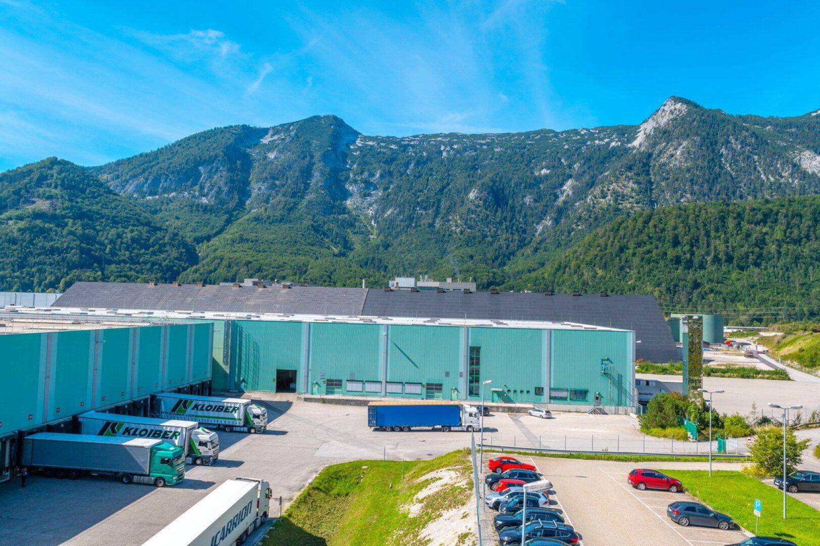 Luftaufnahme eines großen Industriekomplexes mit Laderampen, Parkplätzen und Bergkulisse.
