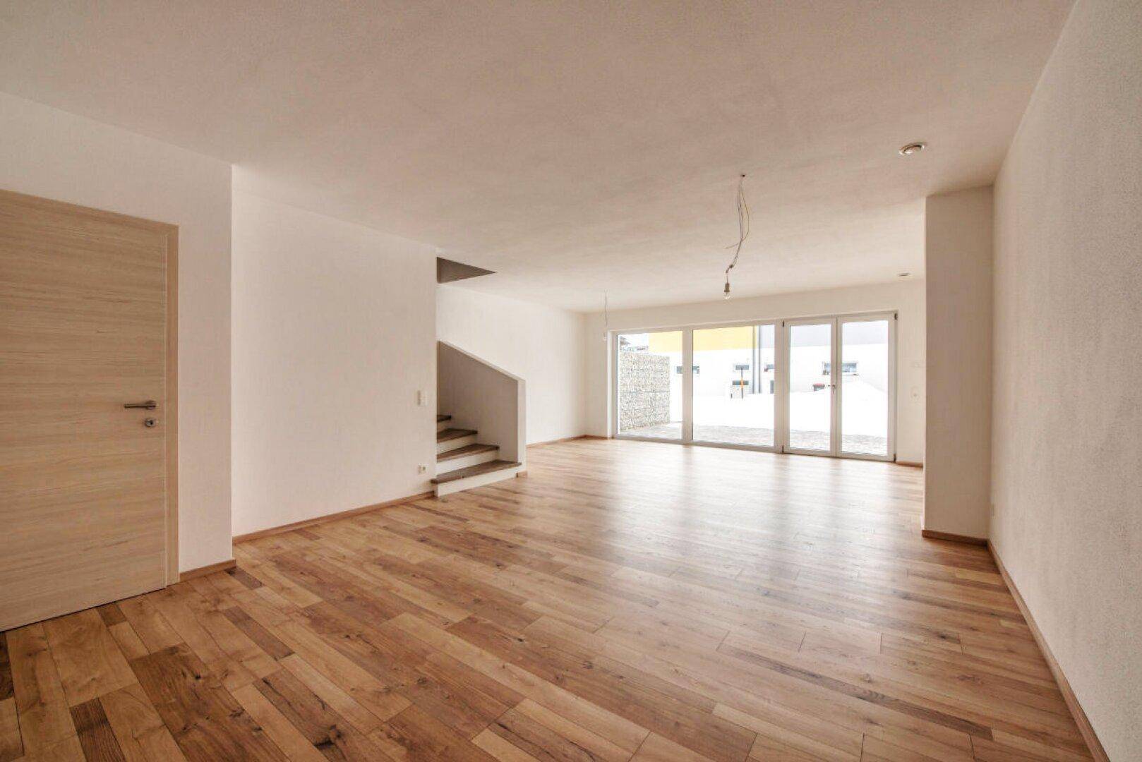 Helles, leeres Wohnzimmer mit Holzboden, großen Fenstern und Zugang zur Terrasse.