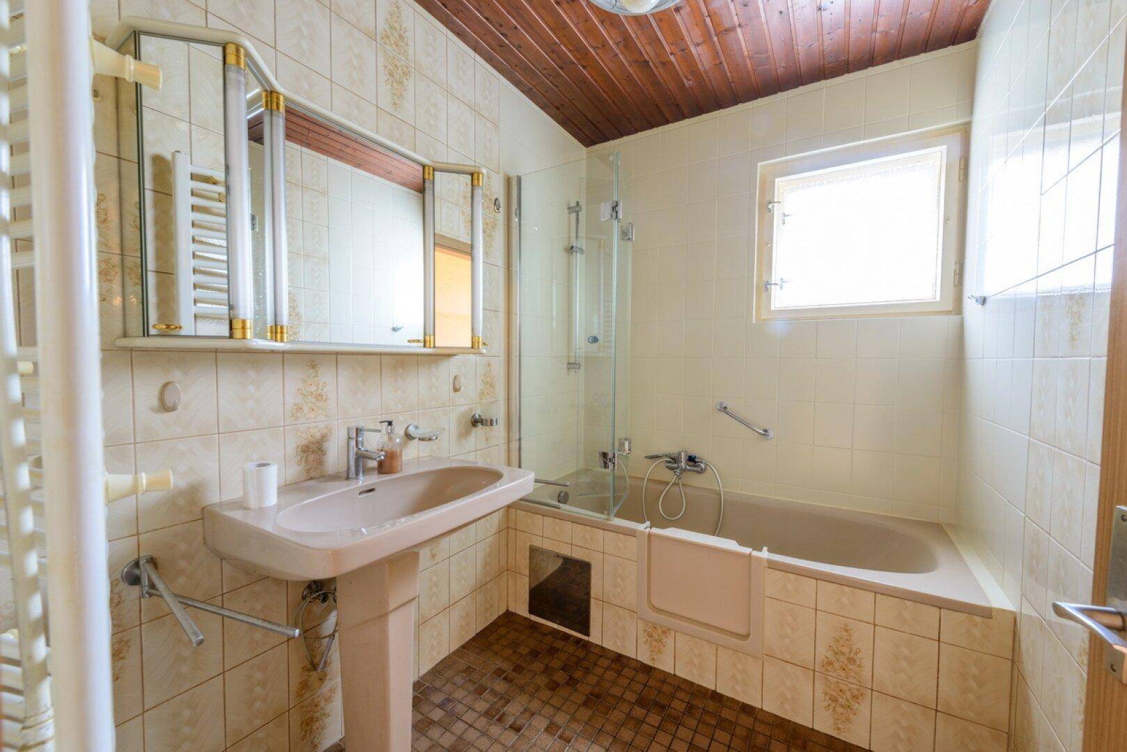 Badezimmer mit Badewanne, Waschbecken und Spiegelschrank, ausgestattet mit beigen Fliesen.
