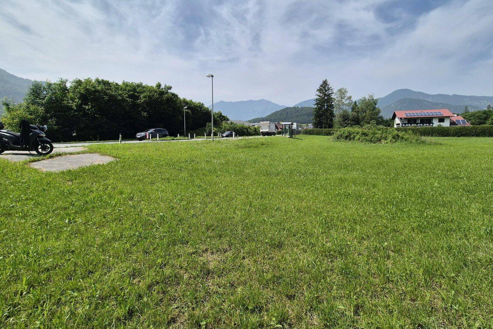 Weitläufige grüne Wiese mit Bergen im Hintergrund und einer Straße am Rand.