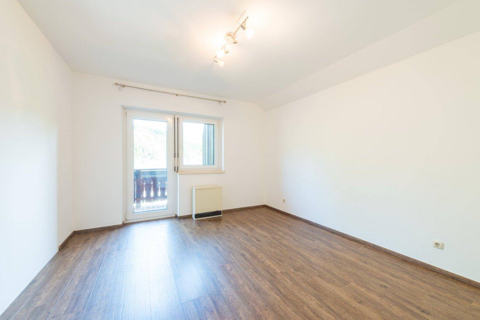 Leeres Zimmer mit Laminatboden, weißen Wänden und Zugang zu einem kleinen Balkon.
